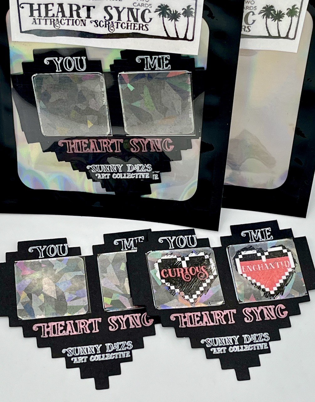 two heart shapred cards with two scratch off spots, the spots of one card are scratched to reveal the hearts beneath, one under the word 'you' says curious, the other, under the word 'me' says enchanted.  below the title is stated: heart sync.  sunny d4z3 art collective